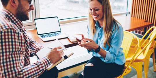 Besprechungssituation Ein Mann mit Notizblock und Stift und eine Frau mit Handy sitzen sich gegenüber und lächeln entspannt. An ihrer Seite steht ein Tisch mit geöffnetem Laptop. Die Stühle sind gelb.